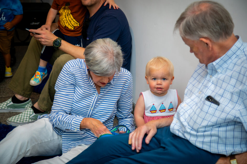 A toddler sits on the floor with two older adults. The woman is looking down at something in her hands, while the man looks at the child, who gazes at the camera. Other people are partially visible in the background.