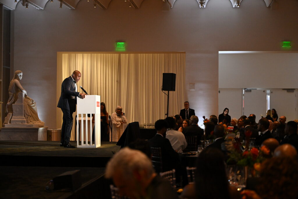 A man speaks at a podium on a stage in front of an audience in a formal, elegant event space with dim lighting and a statue in the background.