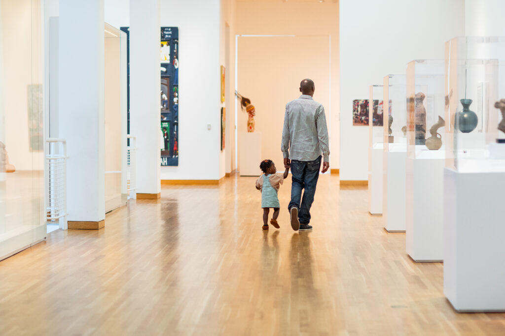 An adult and a child walk hand in hand down a wooden floor hallway in an art museum, surrounded by glass display cases and colorful artwork on the walls.