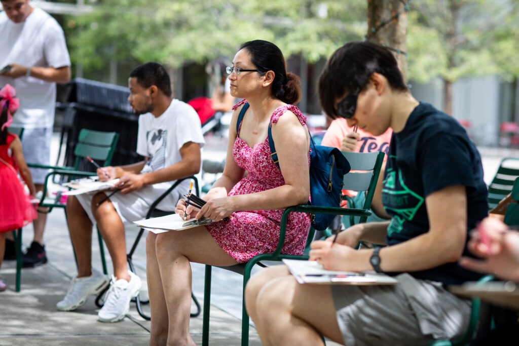 People sit outdoors on green chairs, focused on drawing or writing on clipboards. Trees and other people are visible in the background.