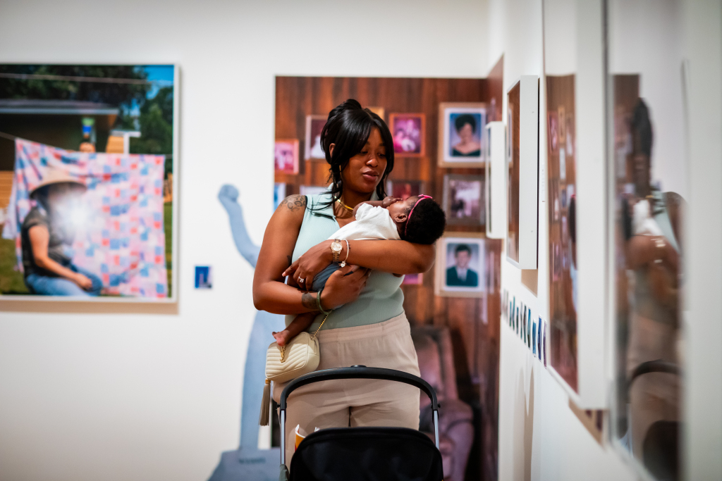 A woman holds a baby while standing in an art gallery, looking at framed photographs on the wall. A stroller is in front of her. Colorful artwork and portraits are visible in the background.