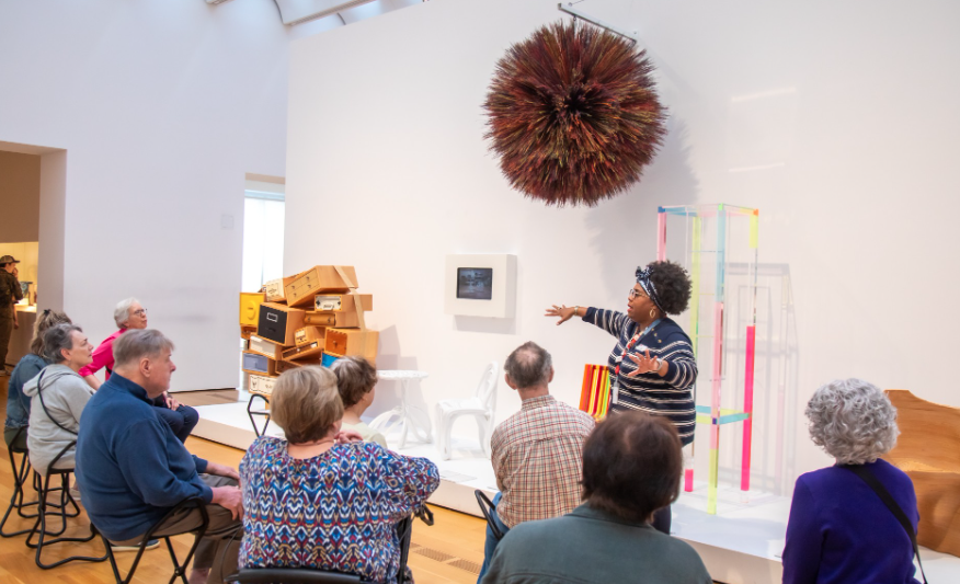 A woman gestures while speaking to a seated group in an art gallery. Colorful sculptures and a spiky hanging artwork are displayed nearby. The attendees listen attentively.