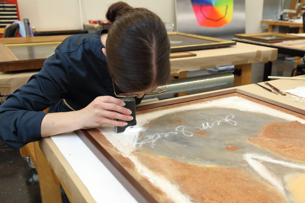 A conservator uses a handheld magnifying glass to examine the surface of an unframed painting in a studio.