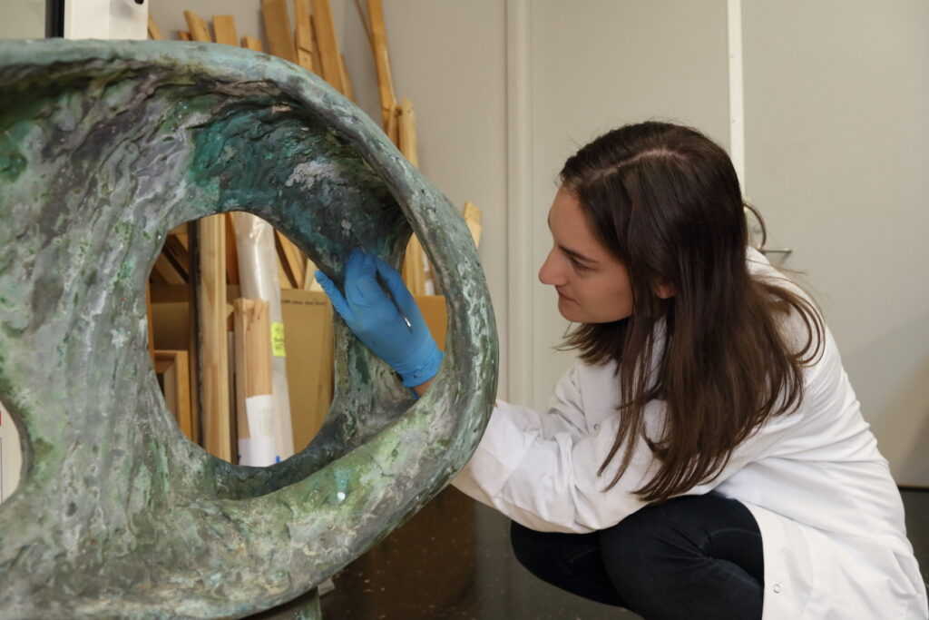 A conservator in a white lab coat and blue gloves uses a small tool to examine a large, weathered bronze sculpture.