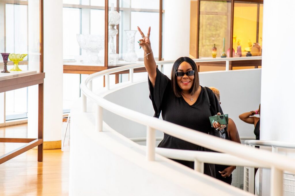 A woman in a black dress and sunglasses makes a peace sign while walking along a curved walkway in a gallery.